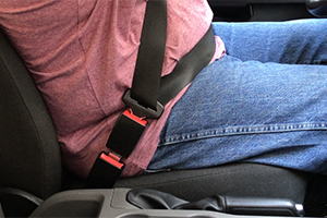 A black, 3” rigid seatbelt extension is buckled in to a seat belt on both ends, securing a man wearing jeans and a red shirt in the driver's seat of his car.